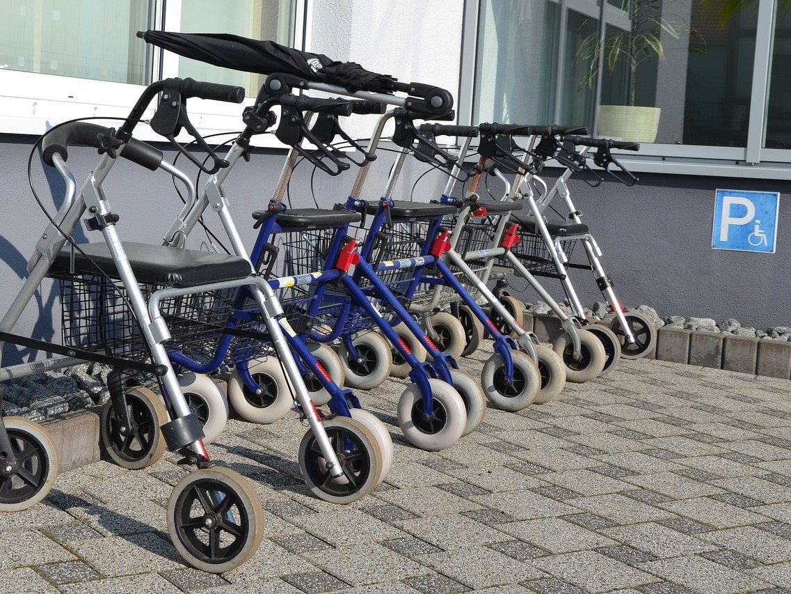 Mehrere Rollatoren stehen auf einem "Parkplatz" für Rollatoren und Rollstühle vor einem Gebäude.