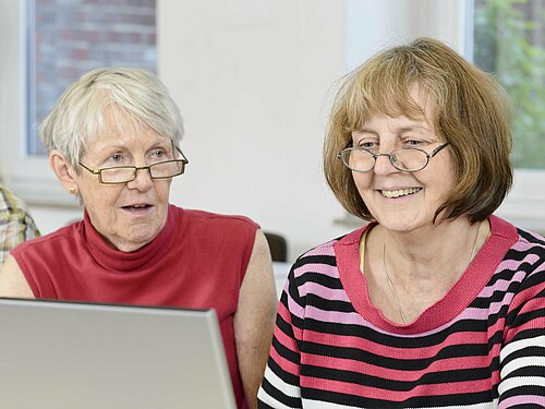 zwei lächelnde Damen sitzen vor einem Laptop und informieren sich über Arthrose