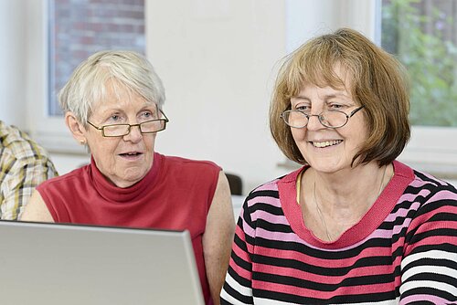 zwei lächelnde Damen sitzen vor einem Laptop und informieren sich über Arthrose