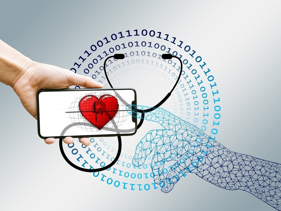 Symbolillustration Digitalisierung Gesundheitswesen: Eine Hand hält ein Smartphone, auf dem ein Herz zu sehen ist. Im Hintergrund ist ein Stethoskop abgebildet.